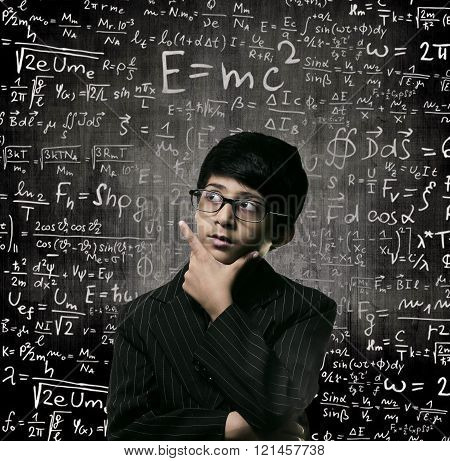 Cute Intelligent Little Boy Looking Up Thinking andWearing Glasses Standing Before A Chalkboard Chemical Formulas Are Written On Board
