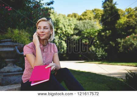 Hipster girl with good mood is calling via cell telephone while sitting in the fresh air in warm spring day gorgeous female is talking with her friend via mobile phone while resting in park outdoors