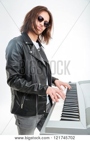 Confident young keyboardist in sunglasses standing and playing on synthesizer over white background