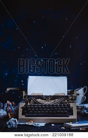 Modern typewriter with sheets of paper, empty coffee cups, pencils and notepads on a dark background. Editing and copywriting workplace. Creative writing concept.