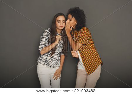 Young woman telling her girlfriend some secret. Two women gossiping. Excited emotional girl whispering to her friend ear, gray studio background
