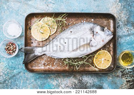 Fresh uncooked dorado or sea bream fish on tray with lemon slices and herbs over blue background, top view