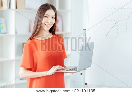 Data base. Merry glad pretty woman standing near white board while gazing  at the camera and  using laptop