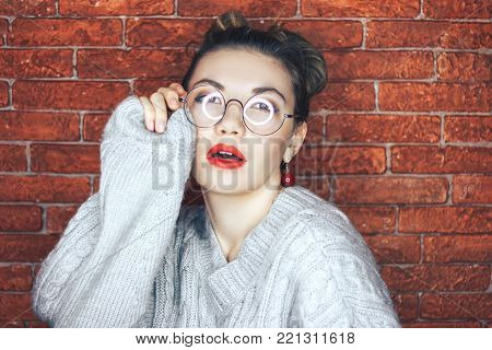Charming brunette young woman in glasses wearing gray sweater touching glasses with eyes closed posing. Red brick background. Red lips.