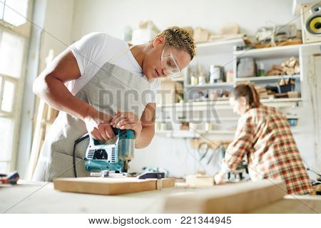 Carpenter in protective eyeglasses sawing wood with electric handtool in workshop