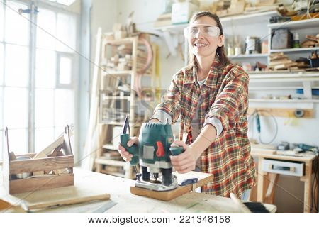 Happy young female carpenter with electric grinder looking at camera while working