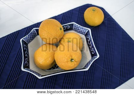 Several Mandarin oranges arranged in an oriental dish on blue place mat on white surface.