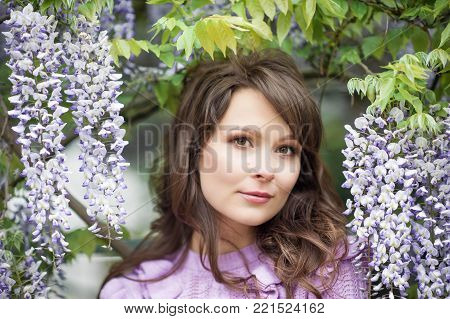 Portrait of a beautiful young brunette woman. A young woman is walking along the spring park. The girl is standing near the wisteria. Blooming park. Spring.