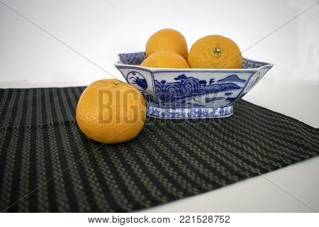 One mandarin orange on striped mat beside an oriental dish filled with oranges.