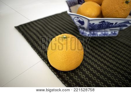 One mandarin orange on a striped place mat in front of dish full of oranges all on a white surface.