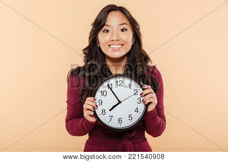 Happy emotions of asian woman with curly long hair holding clock showing nearly 8, waiting for something pleasant over peach background