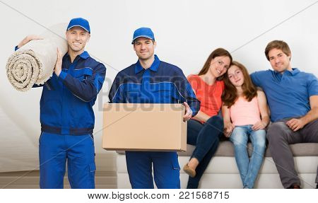 Two Smiling Mover  Men Carrying Cardboard Box And Carpet In Front Of Family