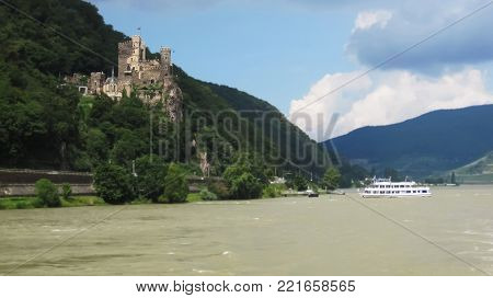Ancient,castle Rheinstein on the rocky cliff along the river Rhine above the green forest while the white turistic ship sail to the castle to embarkart the guests