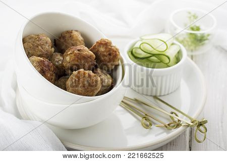 Meat home juicy meatballs in a white ceramic pial on a light background with cucumbers on a side dish. Selective focus