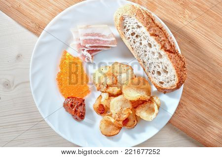 Tasty harmful food. Greasy fried potatoes, bacon, red caviar, spicy sauce and white bread on a white plate. Snacking.