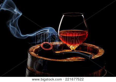 Glass of cognac with smoking pipe on old wooden barrel in front of black background