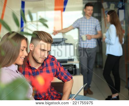 Two confident young people looking at touchpad while their colleagues working in the background