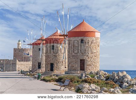 Three Old Windmill On Coast With Blue Cloudy Skay And Lighthouse In The Background