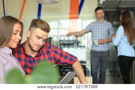 Two confident young people looking at touchpad while their colleagues working in the background