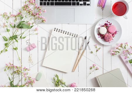 Feminine flat lay workspace with laptop, cup of tea, macarons, flowers, notebook, brushes, on white wooden table. Top view mock up.