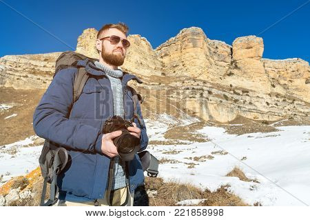 Portrait A bearded hipster photographer with a backpack and wearing sunglasses with a large backpack on his shoulders stands with a DSLR camera in his hands against the background of epic rocks and snow at the foot. The concept of free life in travel and 