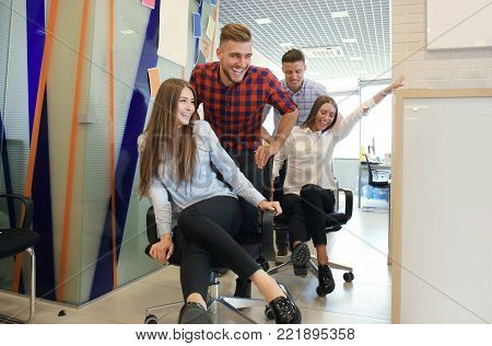 Young cheerful business people in smart casual wear having fun while racing on office chairs and smiling