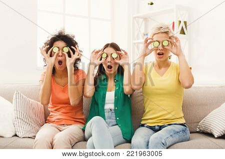 Three beautiful girls in colourful clothes laughing, having fun, covering eyes with cucumber pieces