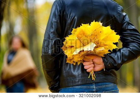 A Guy In A Leather Jacket Holds An Impromptu Bouquet Of Fallen Leaves Behind Him. In Front Of Him In