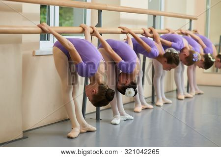 Young Ballerinas Performing Flexibility Exercises. Group Of Young Ballet Dancers Bent Down At Ballet