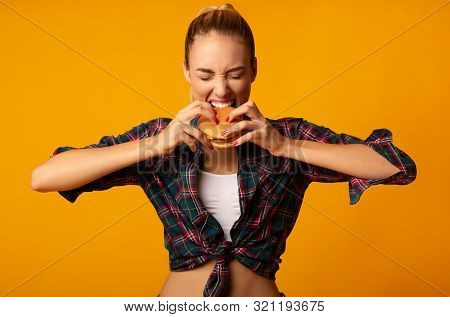 Cheat Meal. Fit Girl Biting Burger On Yellow Studio Background. Young Woman Eating Junk Food.