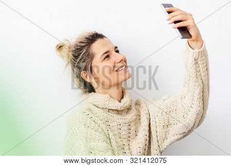 Indoor Image Of Smiling Young Blonde Woman Wearing Knitted Sweater With Hair Bun Making Selfie With 