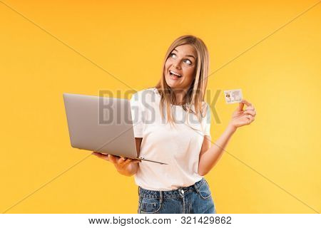 Image of happy blond woman xpressing wonder while holding laptop computer and credit card isolated over yellow background in studio