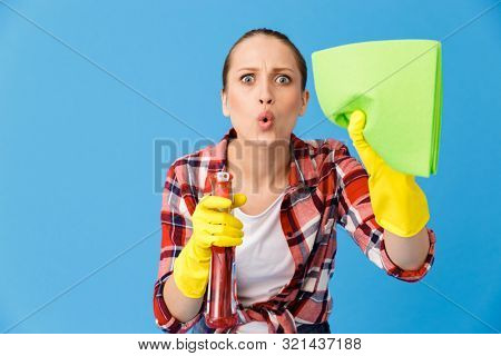 Portrait of uptight housewife in yellow rubber gloves holding detergent sprayer and cleaning rag while doing housework isolated over blue background