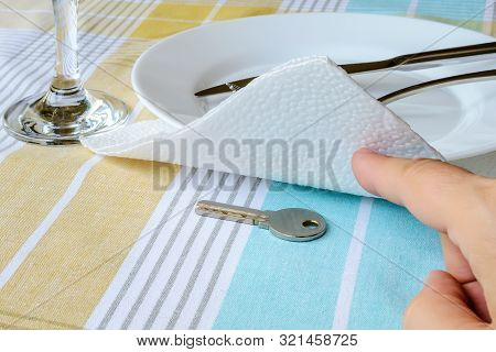 Woman Hand Shows The Key Under The Paper Napkin Near White Plate On The Dining Table. To Hide, To Fi