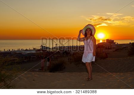 Woman In A White Dress And A Straw Hat On A Background Of The Sea At Sunset.