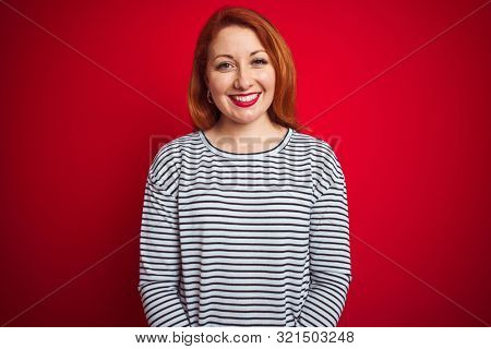 Young redhead woman wearing strapes navy shirt standing over red isolated background with a happy and cool smile on face. Lucky person.
