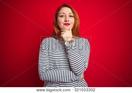 Young redhead woman wearing strapes navy shirt standing over red isolated background looking confident at the camera with smile with crossed arms and hand raised on chin. Thinking positive.