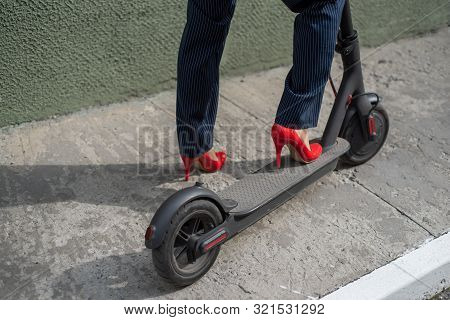 Young Woman In Formal Wear On Red Hight Heels Is Standing On Electrical Scooter. Close-up Of Female 