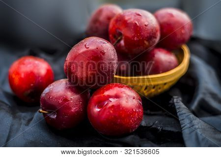 Raw Organic Red Ripe Plums In A Brown-colored Basket On Black Fabric On The Wooden Surface.