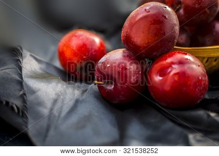 Raw Organic Red Ripe Plums In A Brown-colored Basket On Black Fabric On The Wooden Surface.