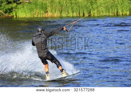 A Wakeboarder Rushes Through The Water At High Speed Along The Banks Of The River, Holding The Cable