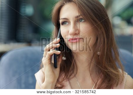 Beautiful Young Brunette Woman In A Pink Dress Is Sitting In A Blue Armchair In A Cafe And Talking O