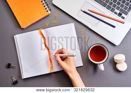 Female Hand Holding Pen And Writing On Notebook During Study Or Planning. Gray Desktop With Orange N