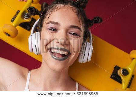 Image of optimistic punk girl with bizarre hairstyle and dark lipstick holding skateboard while listening to music with headphones isolated over red background