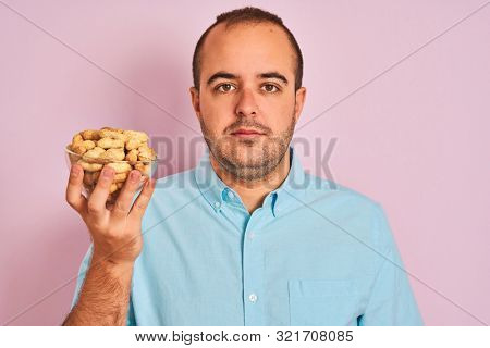 Young man holding bowl with peanuts standing over isolated pink background with a confident expression on smart face thinking serious