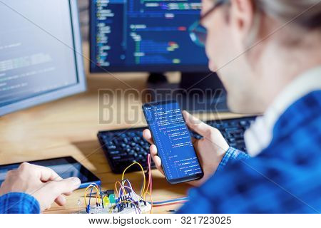 Developer Is Connecting Breadboard To Microcontroller. Man Is Holding Smartphone With Program Code S