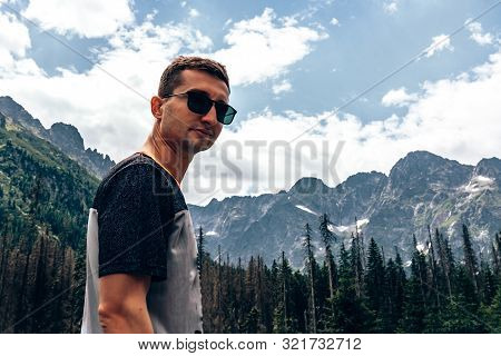 Young Attractive Man In Black Sunglasses And Casual T-shirt Looking At The Camera, Beautiful Mountai