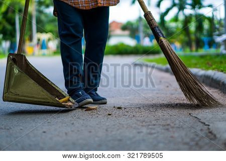 An Asian Woman Is Sweeping Dry Leaves By The Road In The Outdoor Garden. Cleaning Concept.