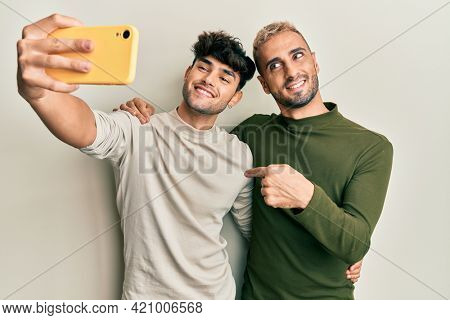 Homosexual gay couple standing together taking a selfie photo with smartphone pointing finger to one self smiling happy and proud 