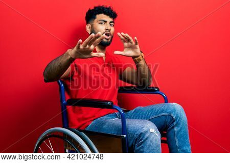 Arab man with beard sitting on wheelchair afraid and terrified with fear expression stop gesture with hands, shouting in shock. panic concept. 
